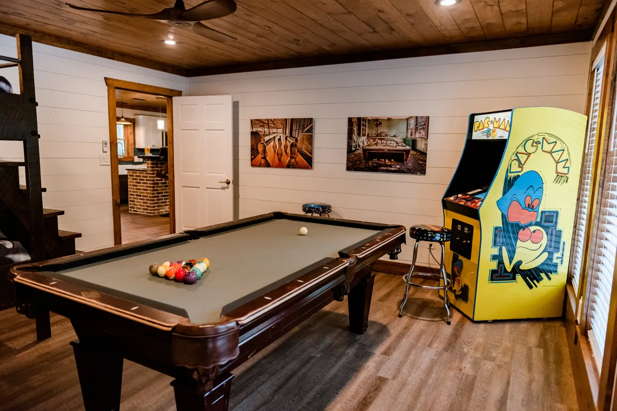 A vibrant game room featuring a pool table and a classic Pac-Man arcade machine, with colorful wall art adding to the energetic atmosphere. The space is arranged for group play, with open space for friends to gather and socialize.