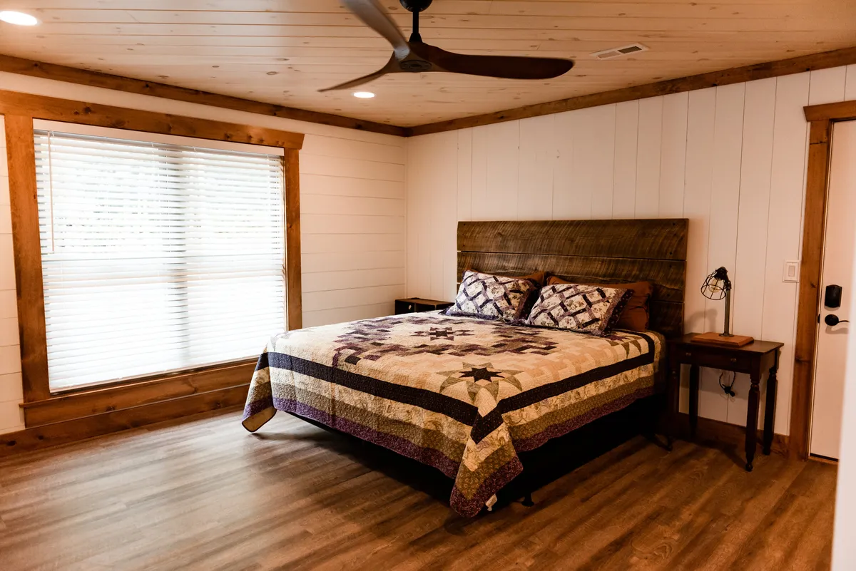 A king bedroom featuring a comfortable bed with a decorative quilt, a large window with blinds allowing natural light to fill the space, and a bedside table with a lamp. The setting is peaceful and inviting, ideal for rest and relaxation after a day of adventure.