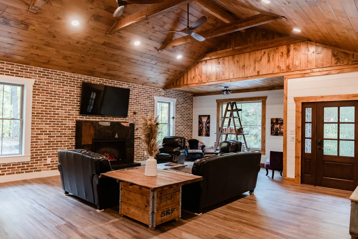 A stylish living room featuring a striking brick wall with a seasonal fireplace as the centerpiece. Plush leather sofas are arranged around a wooden coffee table, and large windows fill the space with views of the surrounding woods. The ambiance is warm and inviting, perfect for evenings of relaxation or sharing stories by the fire.