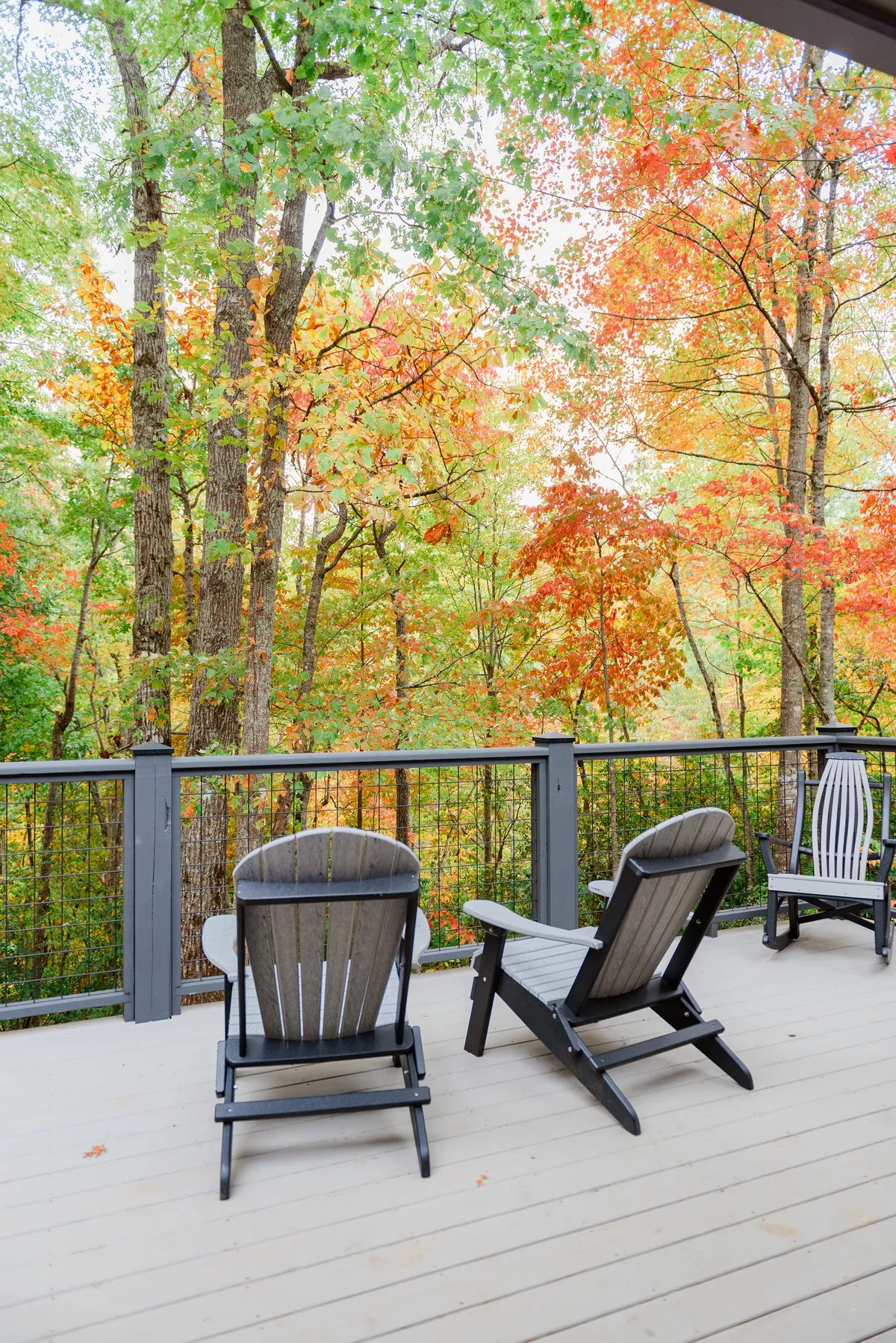This exterior image features a peaceful deck with adirondack chairs overlooking vibrant, colorful foliage. The wooden railing frames a view of the surrounding woods, capturing a tranquil setting ideal for sipping coffee or relaxing in nature.