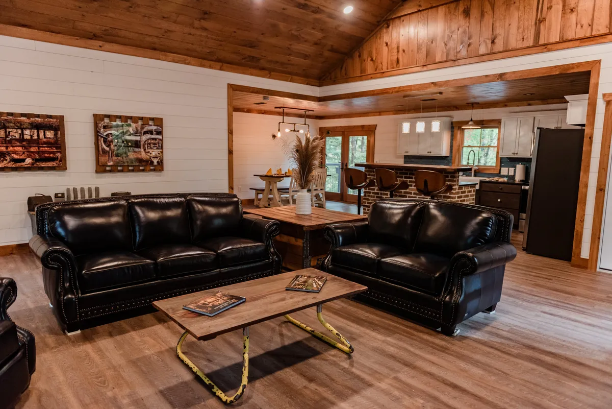 The image features a cozy living area with black leather sofas, a wooden coffee table, decorative wall art, and an adjacent kitchen area. The ambiance is warm and inviting, ideal for relaxing or sharing intimate moments. The space is softly lit, enhancing the sense of comfort and seclusion.