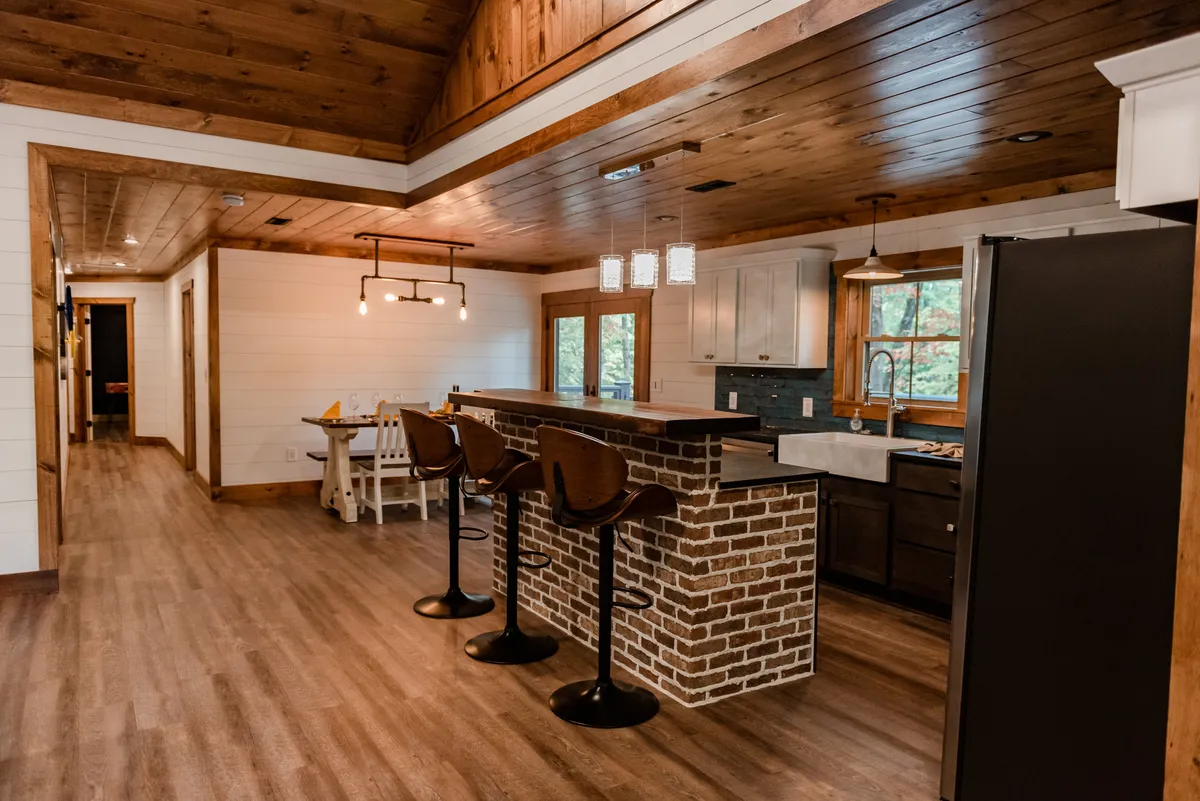 A wide shot of the open-plan kitchen and dining area, showcasing a brick bar counter, modern kitchen appliances, a wooden ceiling, and a dining table ready for meals and gatherings. The space is filled with warm lighting, inviting communal activity.