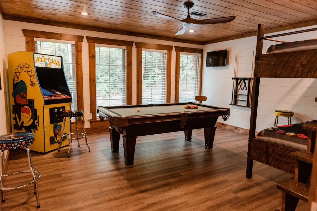 A vibrant game room featuring a pool table at the center, a classic Pac-Man arcade machine against the wall, and a cozy full-over-full bunk bed tucked to the side. The layout emphasizes both recreation and relaxation with ample space for friendly competitions and group hangouts.