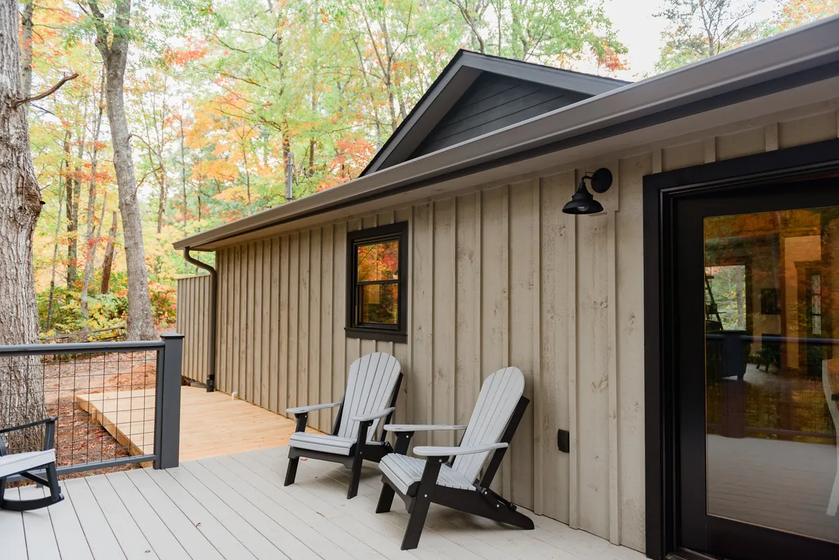 A wooden deck with classic Adirondack chairs is surrounded by mature trees. The setting is peaceful and shaded, ideal for enjoying a morning coffee, reading a book, or simply soaking in the forest view.