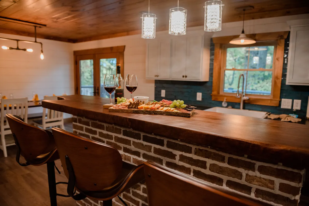 A wooden bar counter set with wine glasses and a charcuterie board forms the focal point, illuminated by hanging light fixtures that create an intimate mood. The setting exudes warmth, ideal for sharing wine and conversation in a romantic retreat.