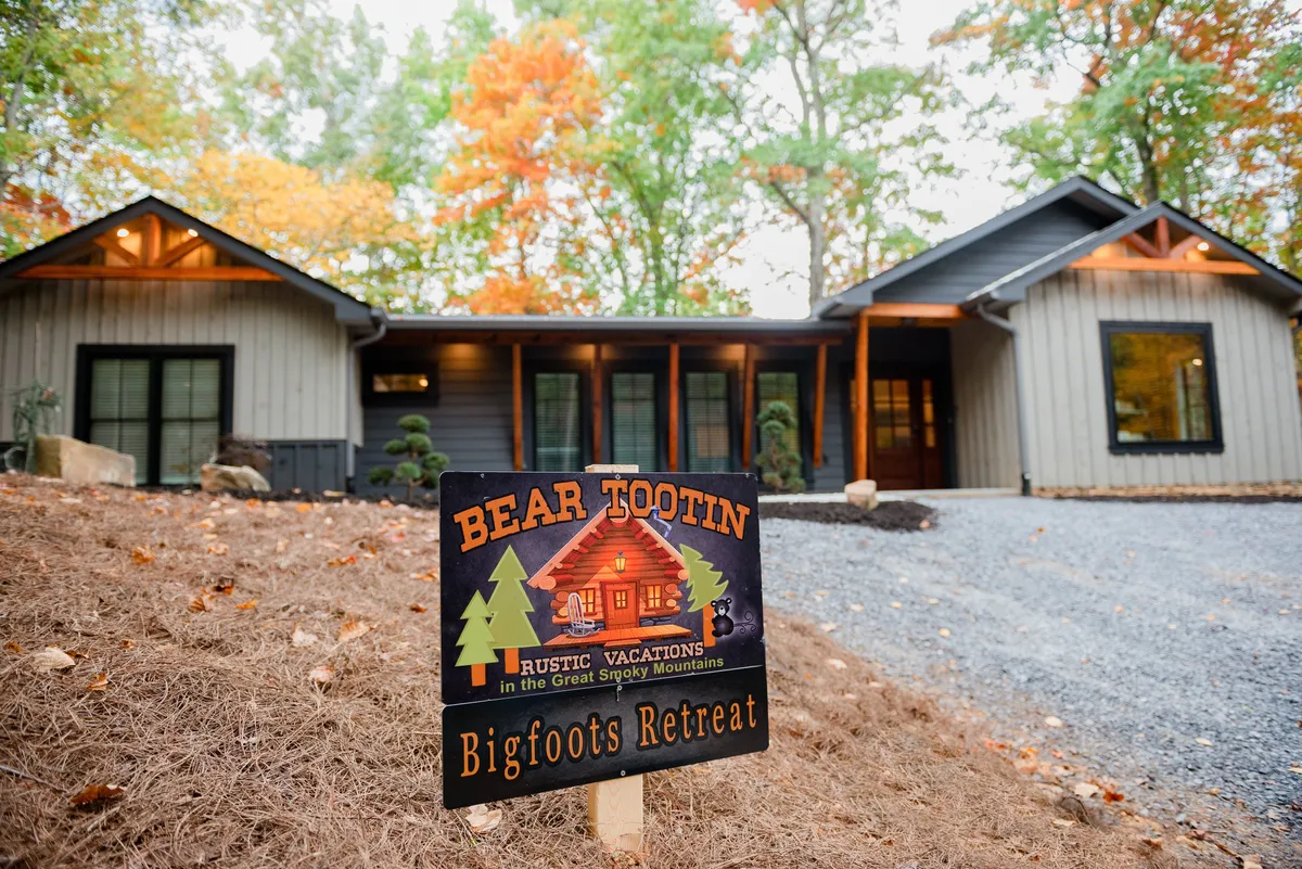 This exterior image captures the front of the property, prominently displaying a rustic sign, the house itself with its modern cabin aesthetic, and vibrant autumn foliage framing the scene. The environment immediately evokes a welcoming, adventurous atmosphere ideal for family gatherings and exploration.